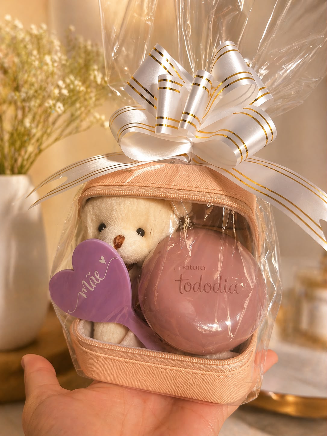 Kit Dia das Mães: necessaire rosé aberta com ursinho de pelúcia, espelho de mão lilás em formato de coração com a palavra Mãe gravada e um sabonete, embalada em celofane com laço branco e dourado.